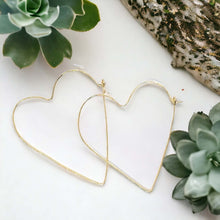 Load image into Gallery viewer, A pair of gold-filled, heart-shaped hoop earrings placed on a white surface with a green plant in the background.