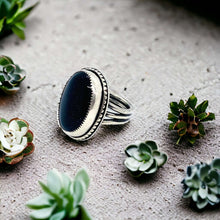 Load image into Gallery viewer, A sterling silver ring with a blue goldstone set in a silver band, displayed on a textured surface with potted plants in the background.